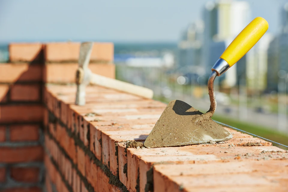 Chantier de construction extérieur avec maçon utilisant une truelle à manche jaune pour appliquer du mortier sur un mur de briques rouges en cours d'édification